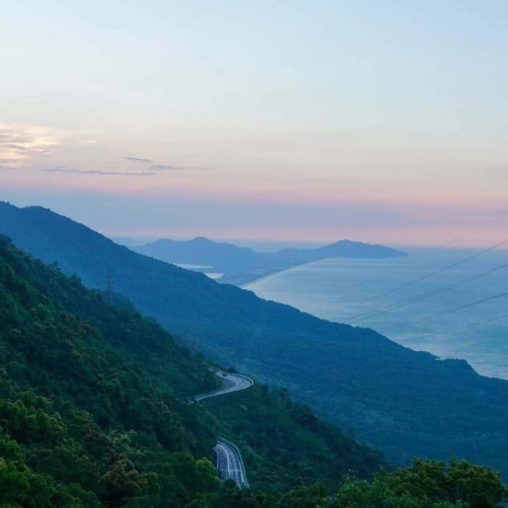 A winding coastal road descending through forested mountains toward the sea at dawn on the Hai Van Pass, Vietnam