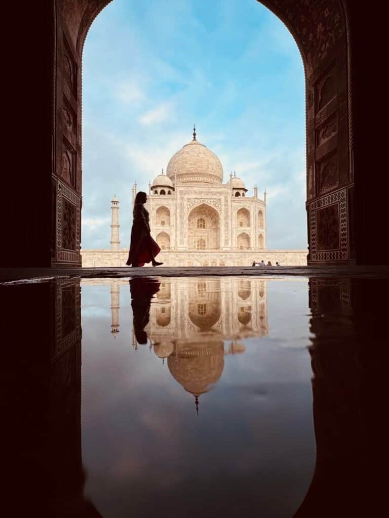 Silhouette of a traveler walking past the Taj Mahal at dawn, framed through an archway and mirrored in still water, Agra, India
