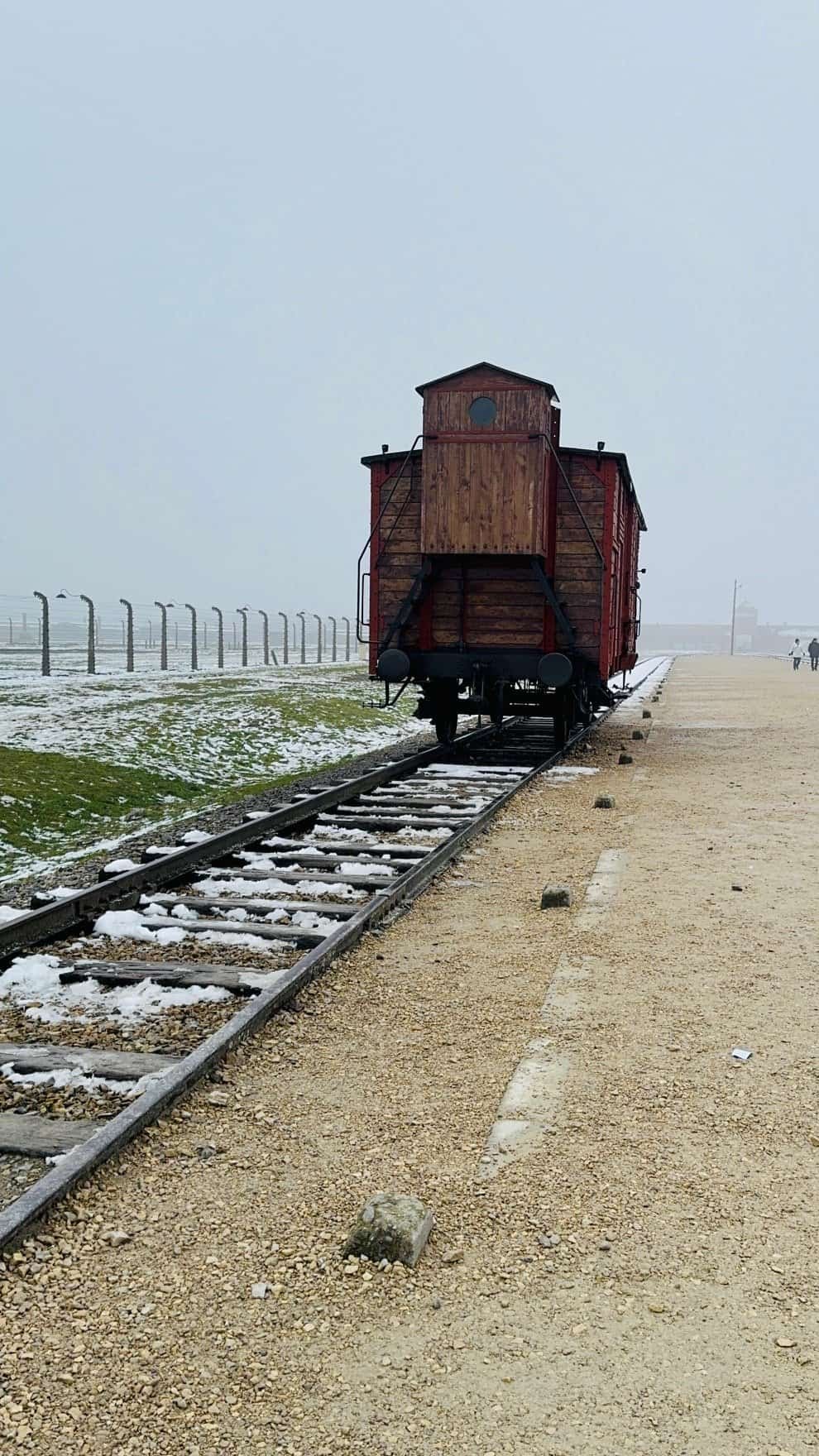 A single wooden boxcar on the tracks at Birkenau, the kind used to transport prisoners, with watchtower fence and gatehouse in the fog