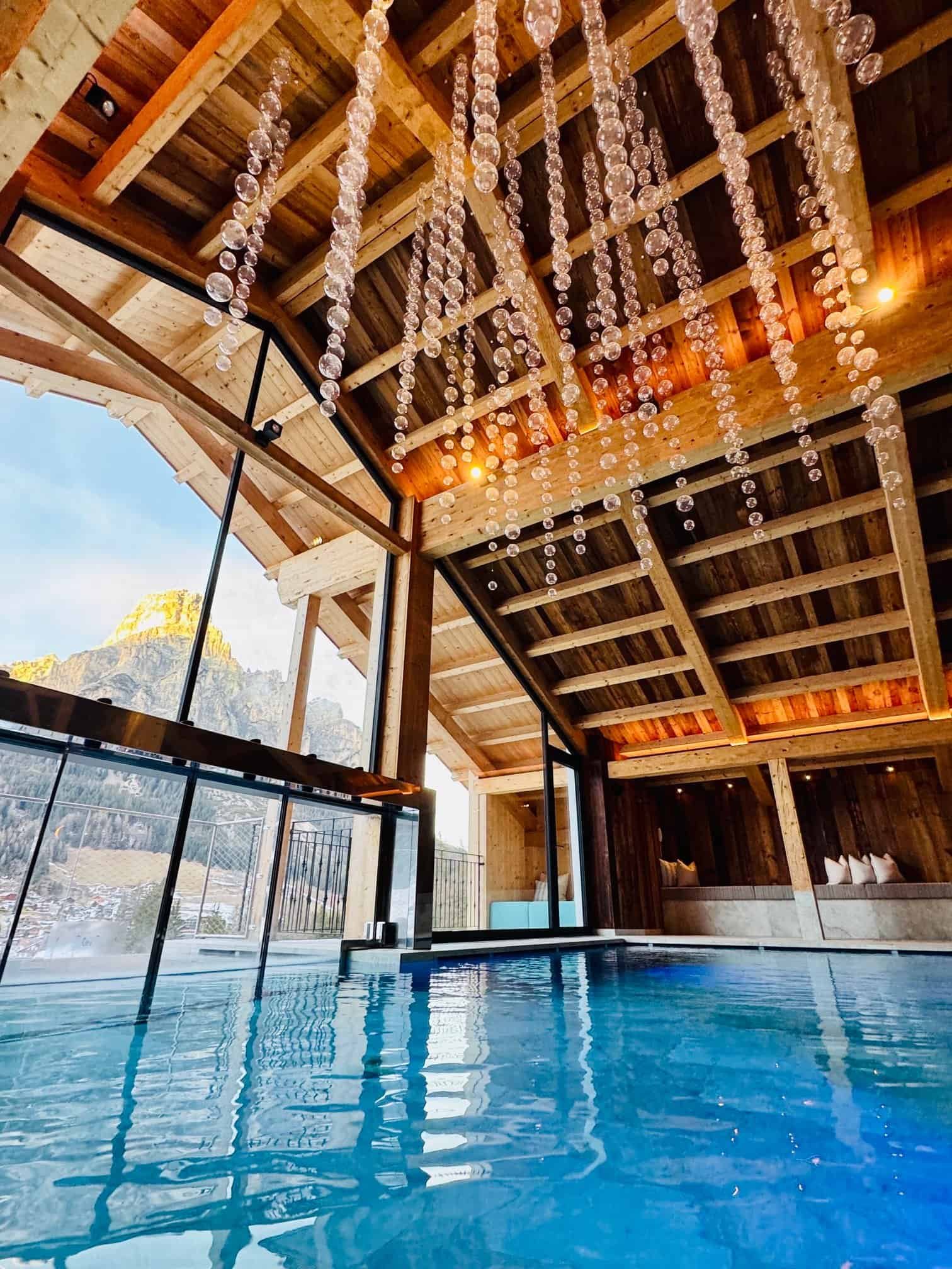 Hotel Arkadia heated pool, hundreds of crystal spheres hanging from exposed timber beams, Dolomite mountain at golden hour through floor-to-ceiling glass