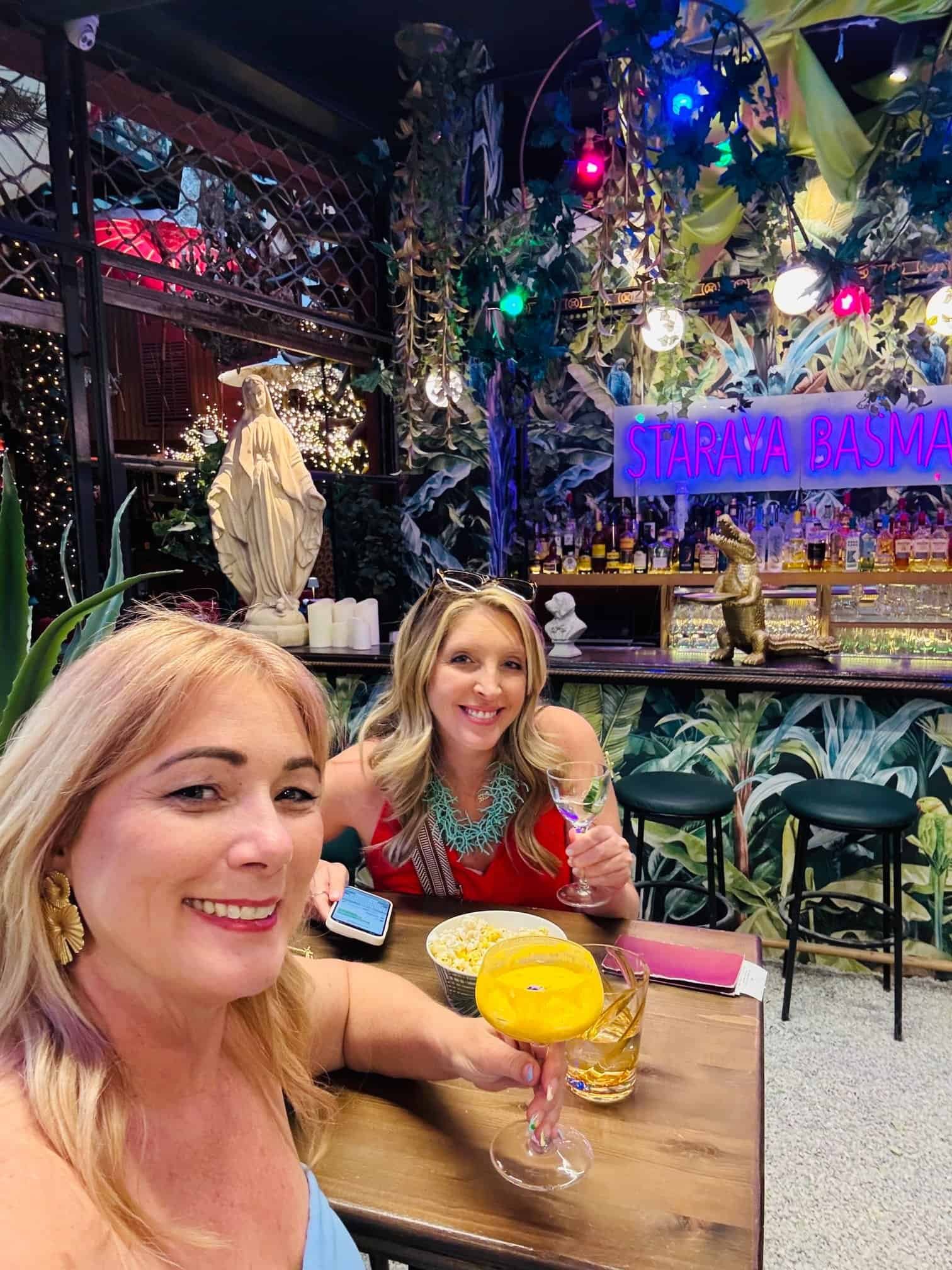 Charity Auger and her friend Lauren with drinks in a lush, plant filled bar in Athens, Greece.