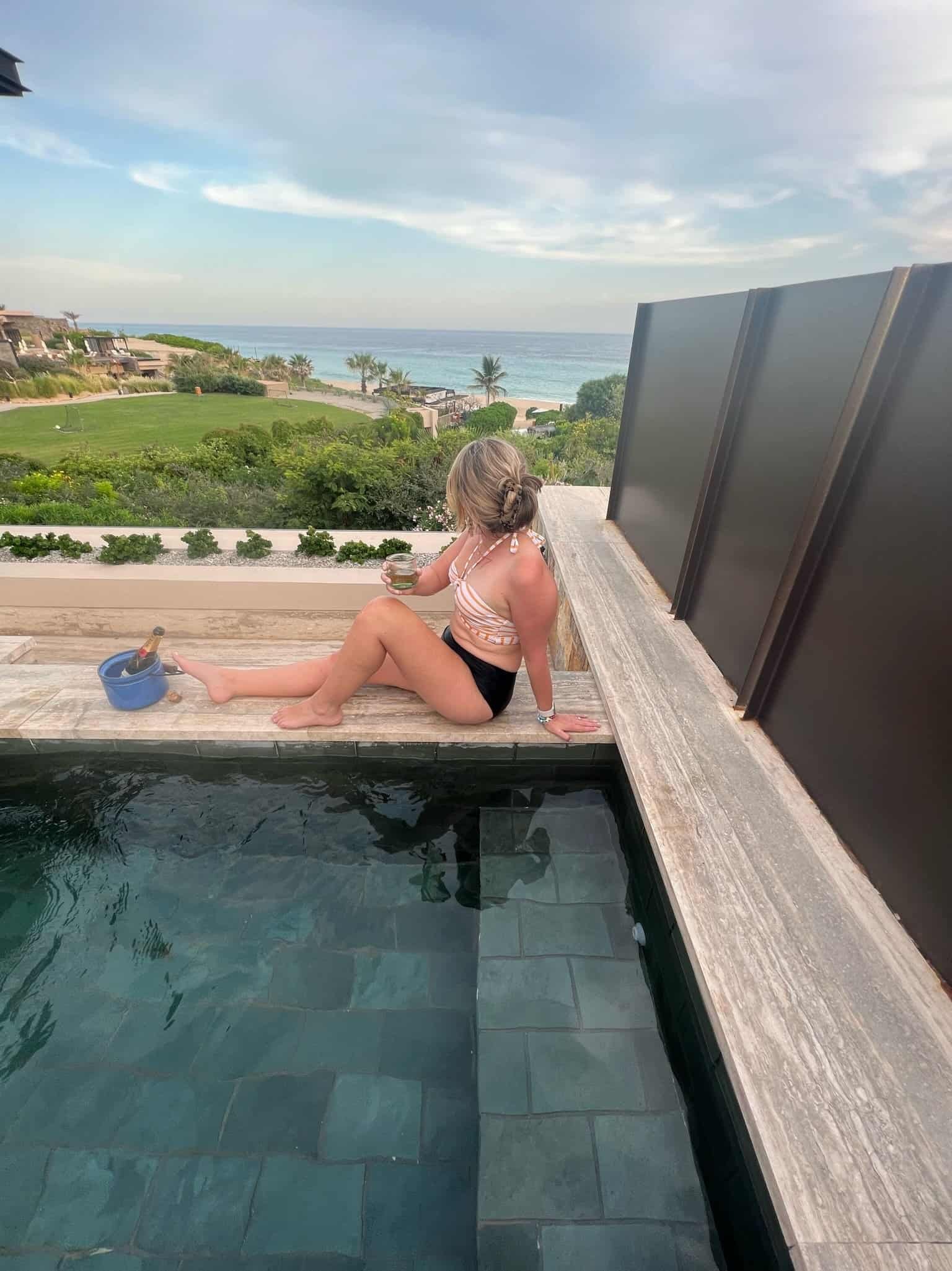 Private pool terrace edge at Zadun, back to camera, cocktail in hand, champagne bucket beside her, Sea of Cortez and resort below
