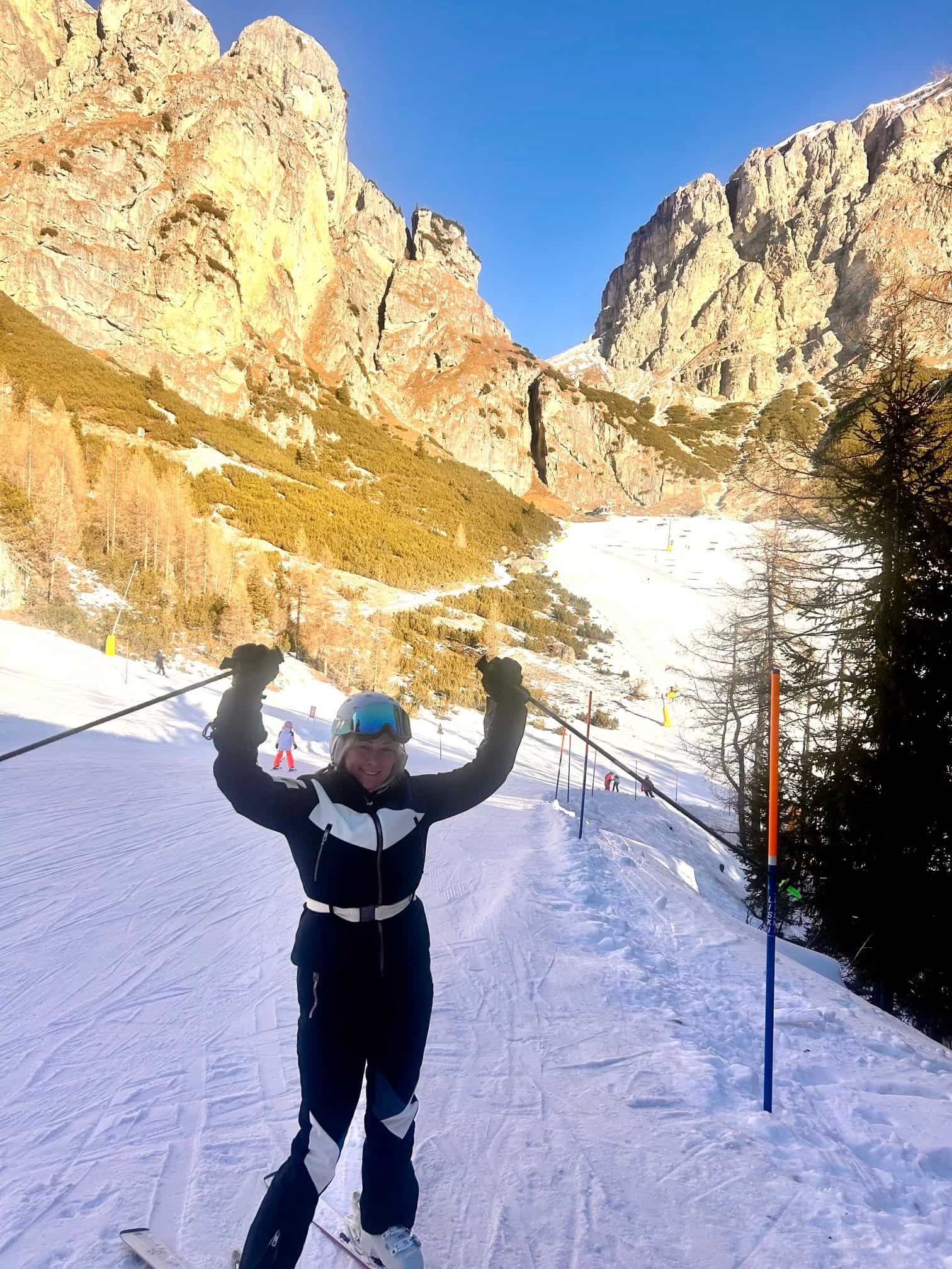 Charity Auger in ski gear with both arms raised on a sunlit run in the Dolomites above Corvara, Italy.