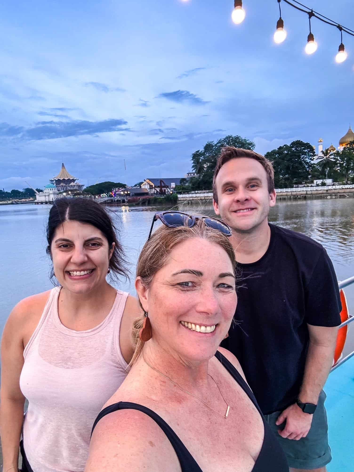 Charity Auger with her friends Josh and Tina at dusk on the Kuching waterfront in Borneo, the golden domes of the city mosque behind them.