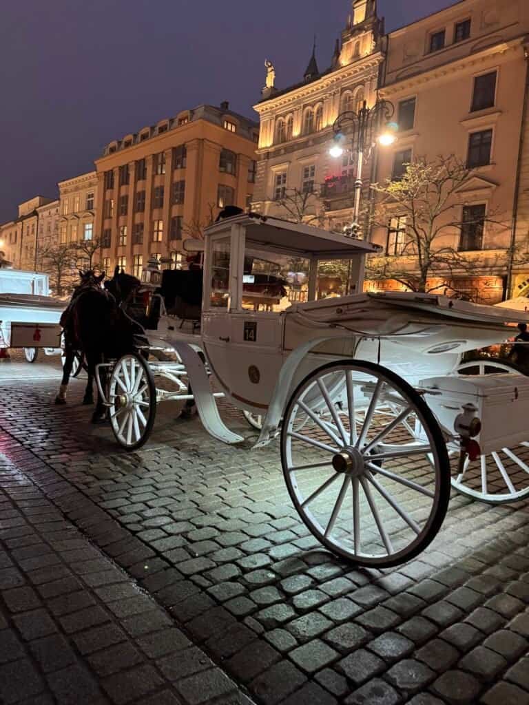 A white horse-drawn carriage on a cobblestone square at night in Krakow, framed by grand illuminated buildings